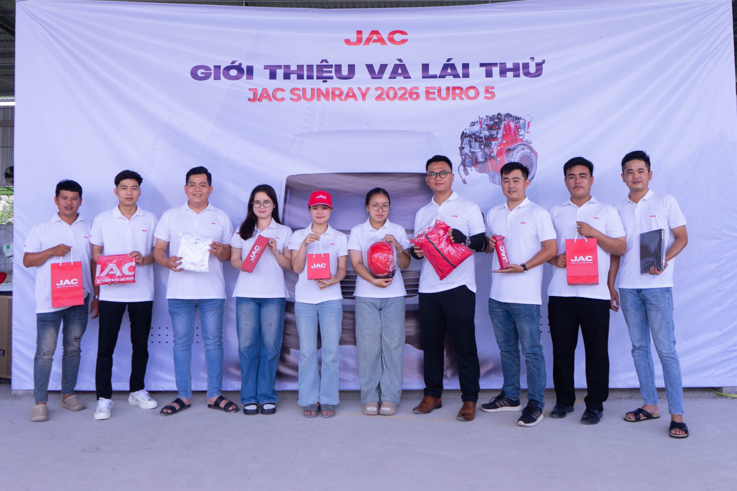 Một nhóm mười người (8 nam, 2 nữ) đang đứng tạo dáng trước một tấm banner lớn màu trắng có logo JAC. Họ đang cầm các vật phẩm quảng cáo như túi, áo, mũ bảo hiểm có thương hiệu JAC. Hầu hết mọi người đều mặc áo polo trắng. Banner có dòng chữ "GIỚI THIỆU VÀ LÁI THỬ JAC SUNRAY 2026 EURO 5" cùng hình ảnh một chiếc xe van màu trắng và một động cơ.
