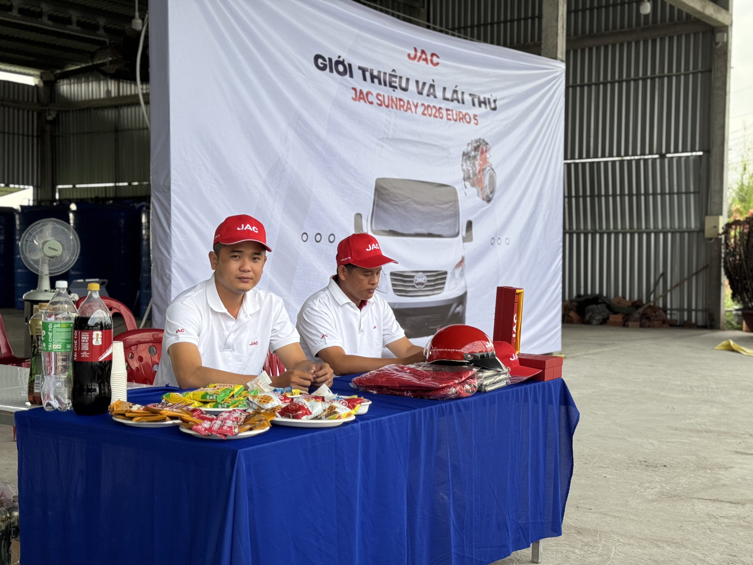 Hai người đàn ông mặc áo polo trắng và mũ lưỡi trai đỏ có logo JAC đang ngồi tại một bàn được phủ khăn màu xanh dương. Trên bàn có đồ ăn nhẹ, đồ uống và các vật phẩm quảng cáo. Tấm banner lớn từ ảnh trước được nhìn thấy ở phía sau.