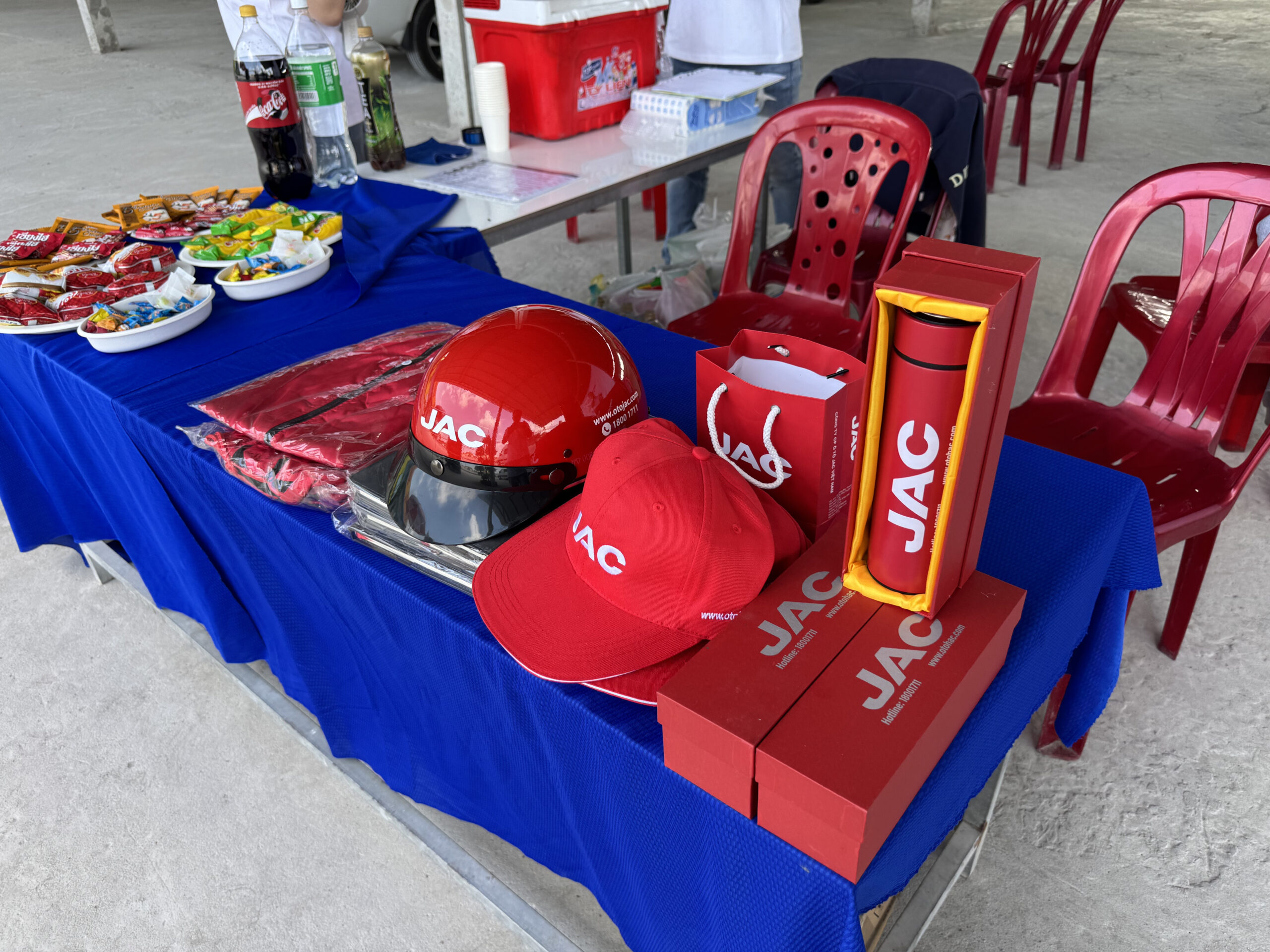 Cận cảnh một bàn được phủ khăn màu xanh dương, trưng bày các vật phẩm quảng cáo của JAC. Bao gồm một mũ bảo hiểm màu đỏ, một mũ lưỡi trai màu đỏ, túi mua sắm màu đỏ, bình giữ nhiệt màu đỏ trong hộp màu vàng, và một số vật phẩm màu đỏ được gấp gọn (có thể là áo). Đồ ăn nhẹ và đồ uống cũng được đặt trên bàn.
