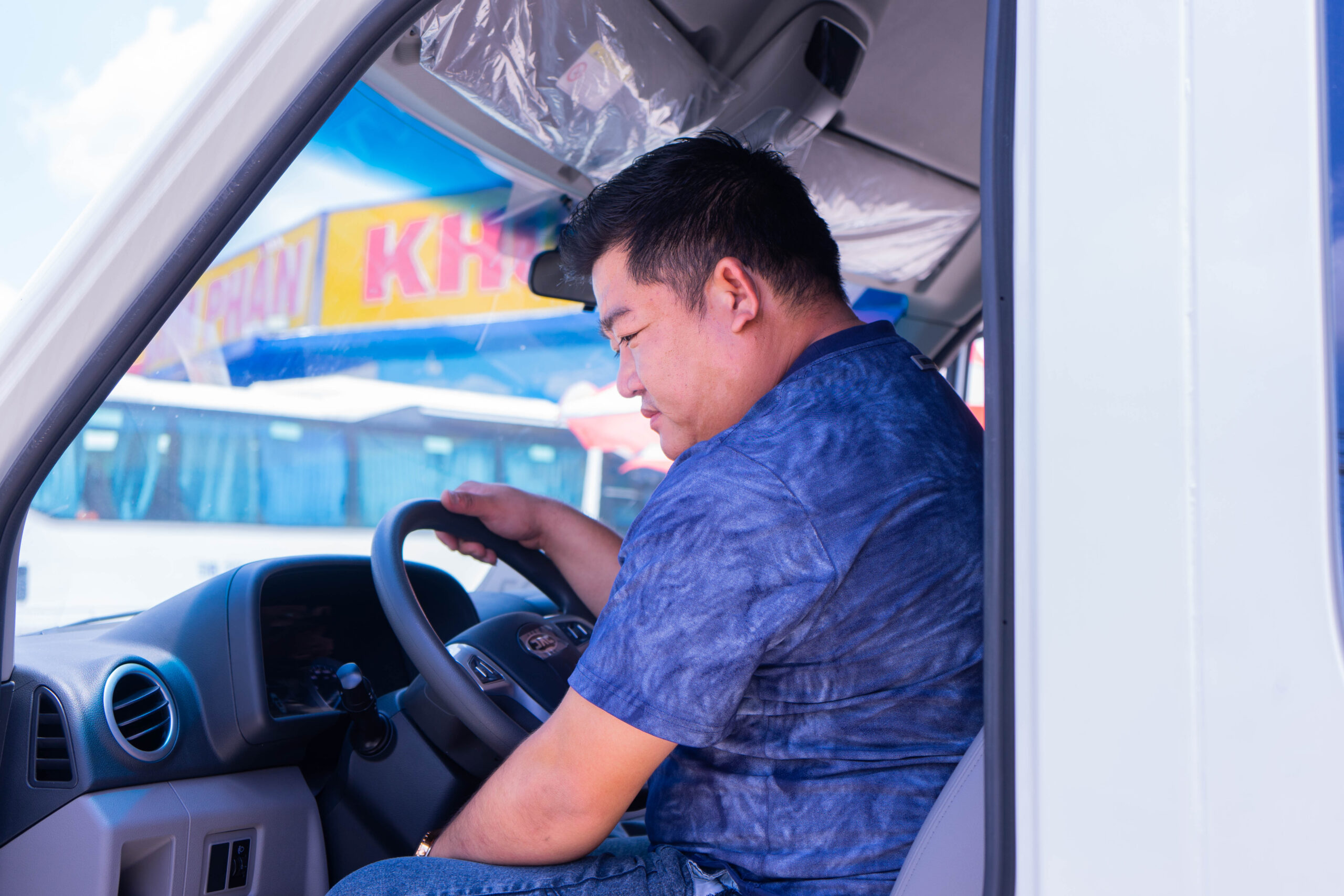 Góc chụp nội thất xe, cho thấy một người đàn ông đang ngồi ở ghế lái của một chiếc xe van màu trắng (có thể là JAC Sunray). Anh ấy đang đặt tay lên vô lăng có logo JAC. Bảng điều khiển và các cửa gió điều hòa cũng được nhìn thấy rõ.