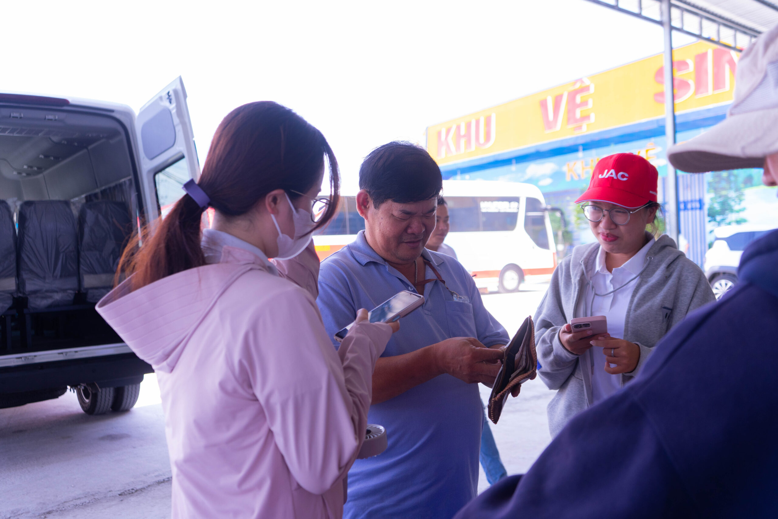 Một nhóm người đang tụ tập và tương tác gần một chiếc xe van màu trắng. Ba người (hai phụ nữ và một người đàn ông) đang nhìn vào điện thoại của họ. Một người đàn ông khác chỉ xuất hiện một phần. Chiếc xe van màu trắng (JAC Sunray) đang mở cửa bên hông ở phía sau.