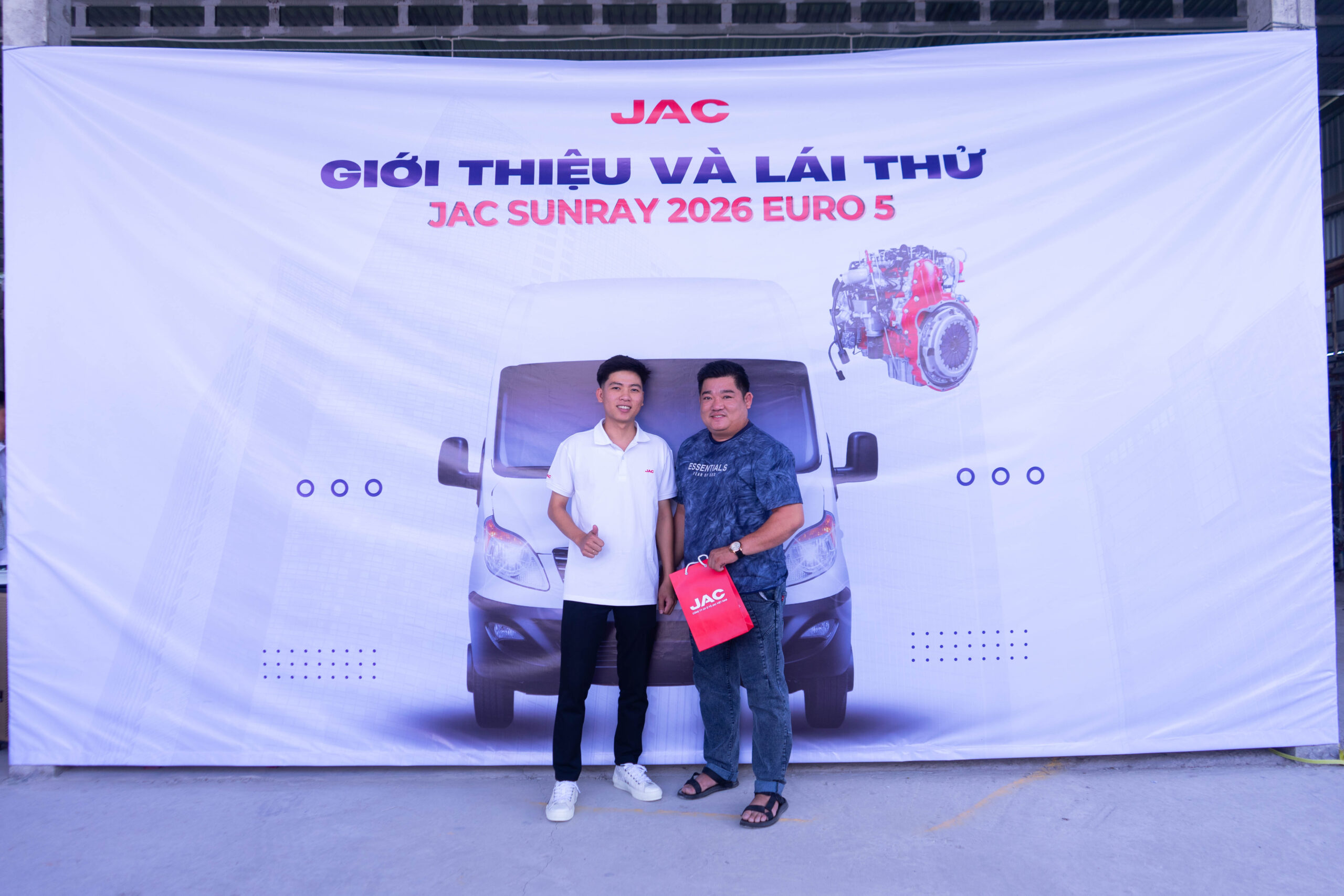 Hai người đàn ông đang tạo dáng chụp ảnh trước tấm banner lớn màu trắng có logo JAC. Một người đàn ông trẻ mặc áo polo trắng và quần đen đang giơ ngón tay cái, đứng cạnh một người đàn ông lớn tuổi hơn mặc áo phông xanh họa tiết và quần jean, đang cầm một chiếc túi đỏ có logo JAC. Tấm banner hiển thị đầy đủ dòng chữ "GIỚI THIỆU VÀ LÁI THỬ JAC SUNRAY 2026 EURO 5" cùng hình ảnh xe van và động cơ.