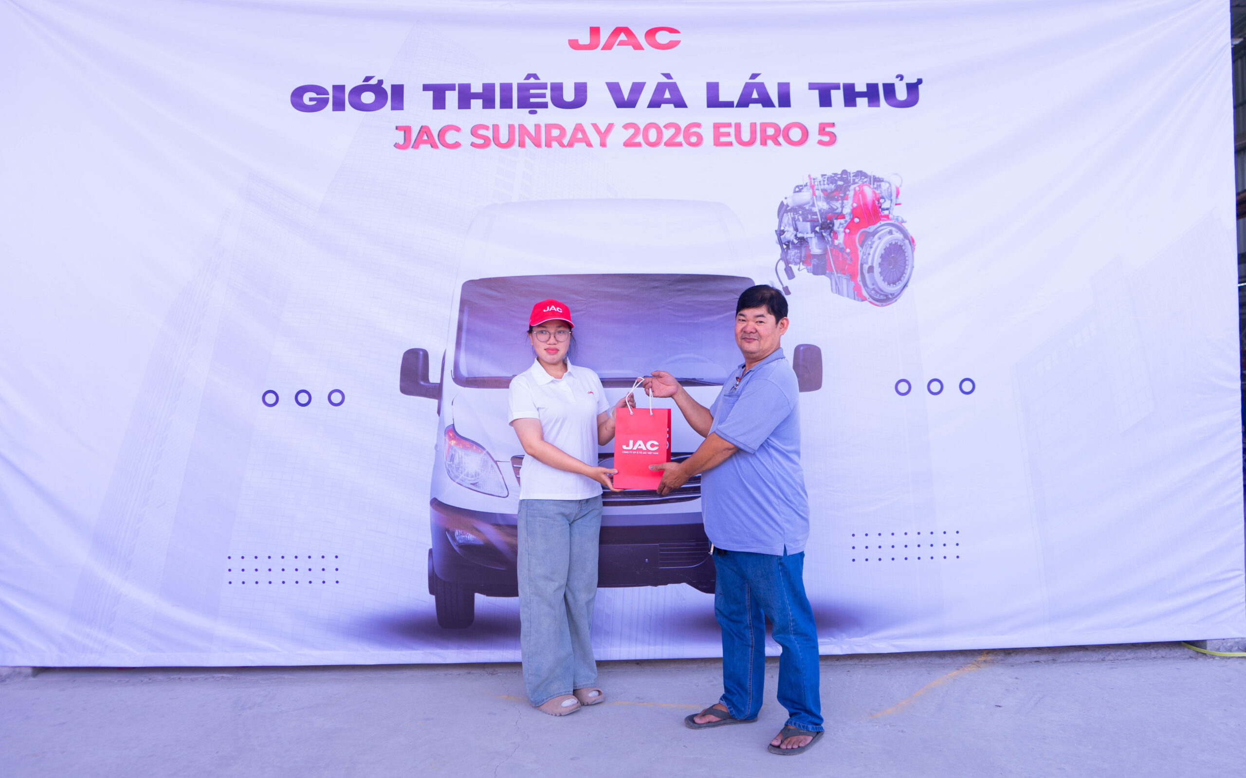 Một phụ nữ và một người đàn ông đang tạo dáng chụp ảnh trước tấm banner lớn màu trắng có logo JAC. Người phụ nữ mặc áo polo trắng, quần sáng màu và đội mũ lưỡi trai đỏ có logo JAC, đang trao một chiếc túi đỏ có logo JAC cho người đàn ông lớn tuổi hơn mặc áo polo xanh nhạt và quần jean. Tấm banner hiển thị đầy đủ dòng chữ "GIỚI THIỆU VÀ LÁI THỬ JAC SUNRAY 2026 EURO 5" cùng hình ảnh xe van và động cơ.