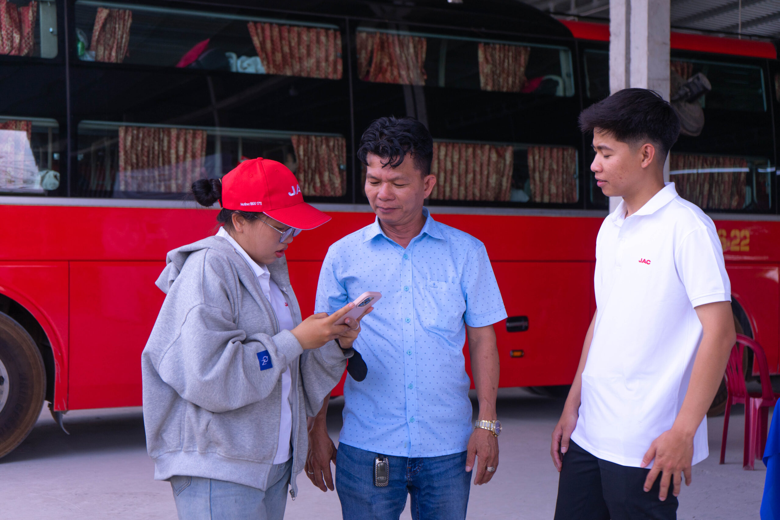 Hai người đang tạo dáng chụp ảnh trước tấm banner lớn màu trắng có logo JAC. Một người đàn ông mặc áo sơ mi xanh họa tiết, đang cầm điện thoại và một chiếc túi đỏ có logo JAC, đứng cạnh một phụ nữ đeo kính, mặc áo polo trắng có logo JAC, đang giơ hai ngón tay cái. Tấm banner hiển thị đầy đủ dòng chữ "GIỚI THIỆU VÀ LÁI THỬ JAC SUNRAY 2026 EURO 5" cùng hình ảnh xe van và động cơ.