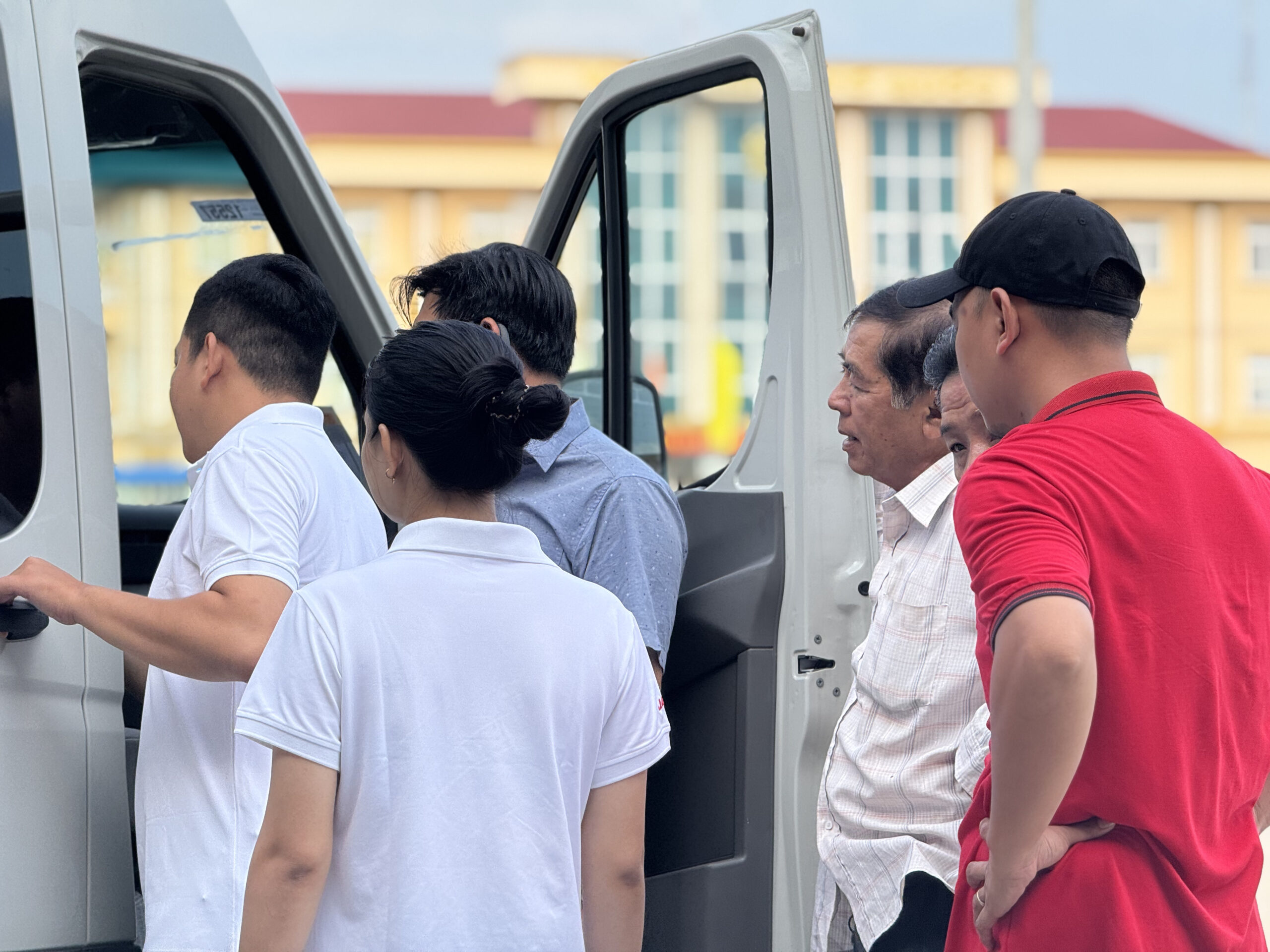 Ba người đang tương tác ngoài trời, với một chiếc xe buýt màu đỏ lớn đậu ở phía sau. Một phụ nữ mặc áo hoodie xám và mũ lưỡi trai đỏ có logo JAC đang cho hai người đàn ông xem gì đó trên điện thoại của cô ấy (một người mặc áo sơ mi xanh nhạt họa tiết, một người mặc áo polo trắng có logo JAC).