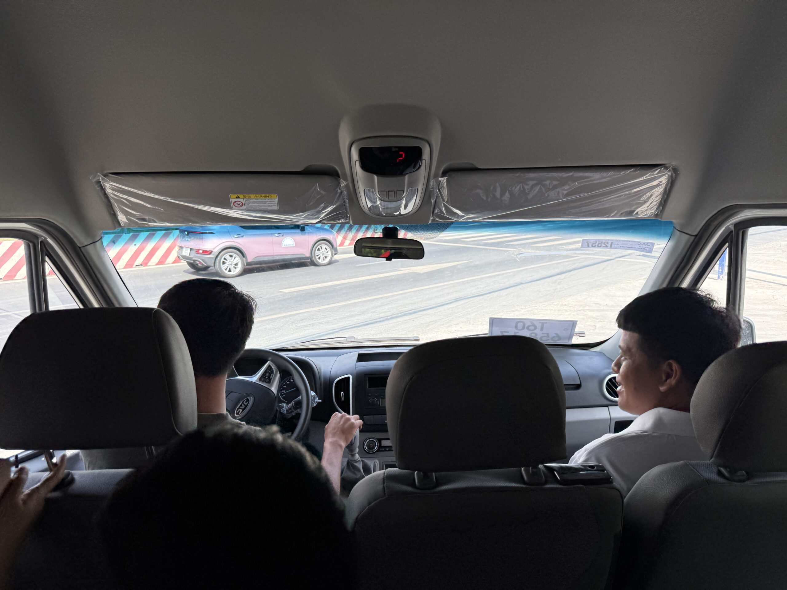 Góc chụp từ ghế lái của một chiếc xe van màu trắng (JAC Sunray). Vô lăng có logo JAC nổi bật. Bảng điều khiển, cụm đồng hồ và các cửa gió điều hòa đều có thể nhìn thấy. Tay của người lái đang đặt trên vô lăng.