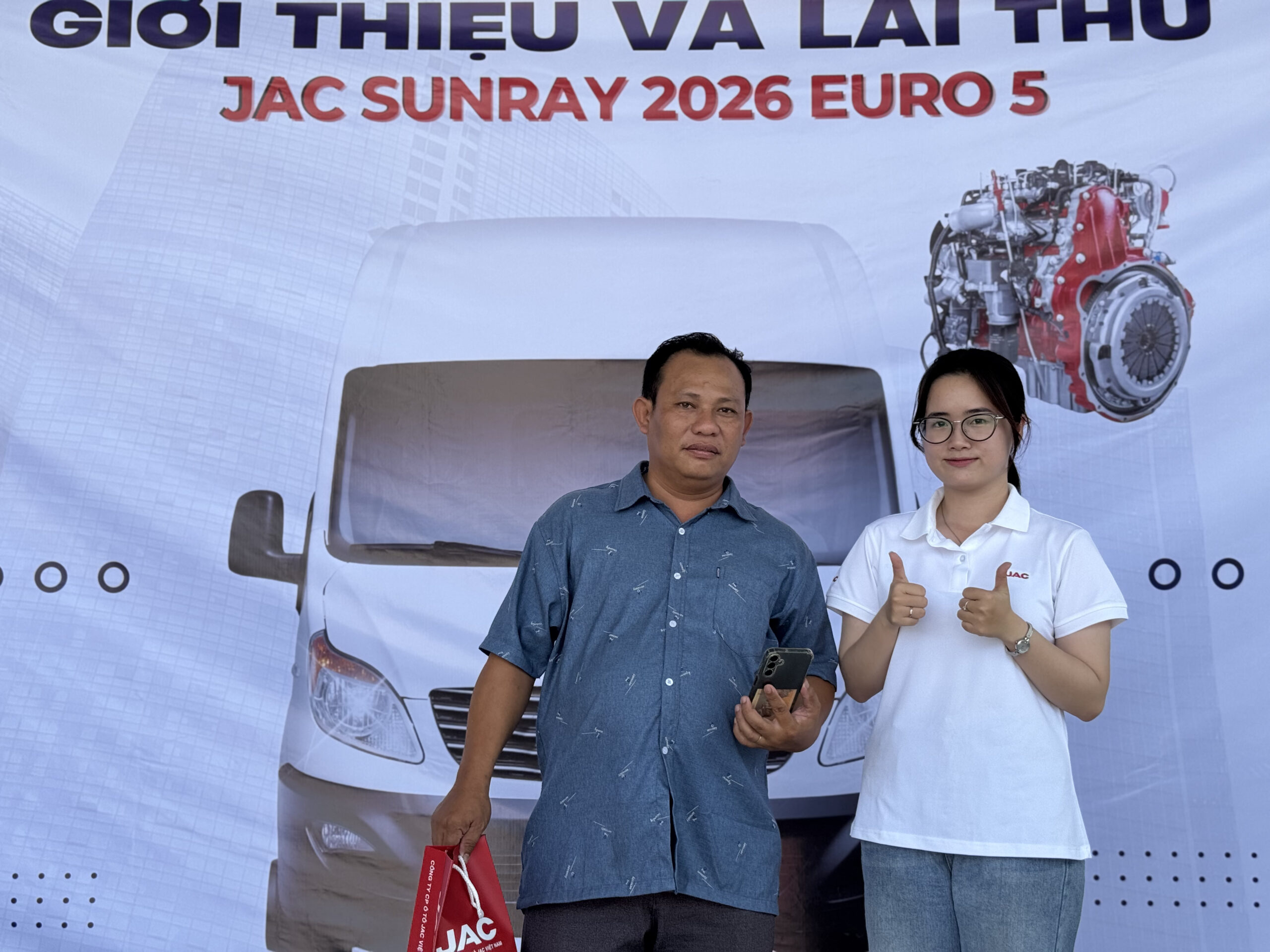 Góc chụp nội thất của một chiếc xe van màu trắng (JAC Sunray) từ hàng ghế sau. Người lái và một hành khách phía trước được nhìn thấy từ phía sau. Con đường phía trước có thể nhìn thấy qua kính chắn gió.