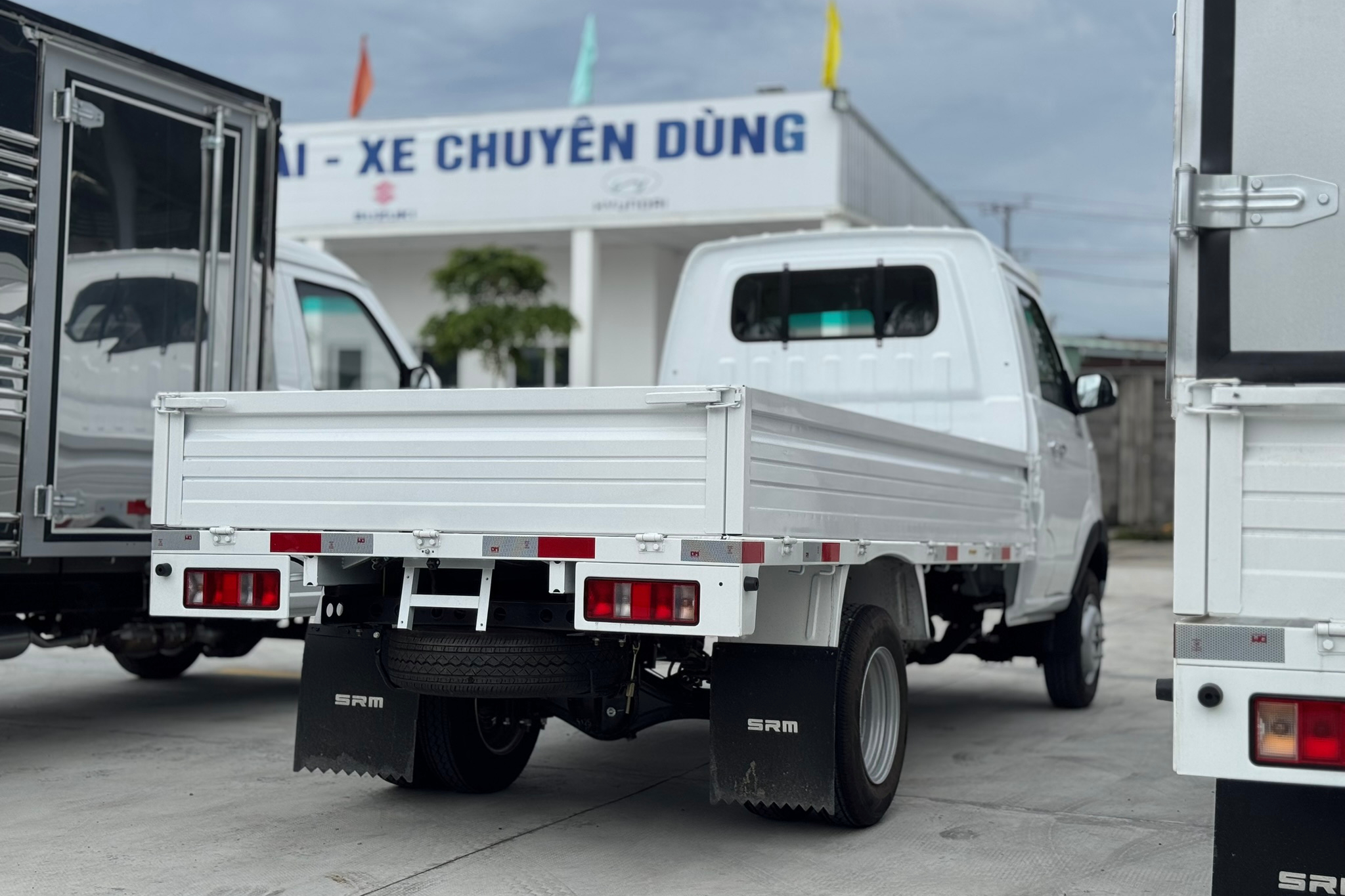 Góc nhìn ba phần tư phía sau của một chiếc xe tải nhỏ SRM màu trắng với thùng lửng. Xe có bánh đôi phía sau, chắn bùn in logo SRM, dải phản quang màu đỏ trên cản sau và lốp dự phòng được gắn dưới gầm. Thùng xe có thành thấp. Một chiếc xe tải thùng kín được nhìn thấy một phần ở bên trái và một chiếc thùng lửng khác ở bên phải. Phía sau là một tòa nhà có chữ 