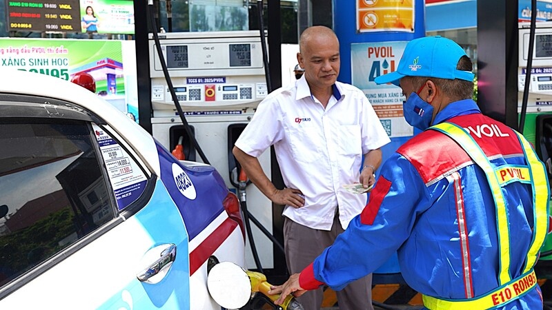 Bối cảnh tại một trạm xăng PVOIL, một người đàn ông mặc áo sơ mi trắng đang thanh toán cho nhân viên PVOIL mặc đồng phục màu xanh. Một chiếc ô tô màu trắng đang được đổ xăng E10 RON95-V. Trụ bơm xăng hiển thị rõ.